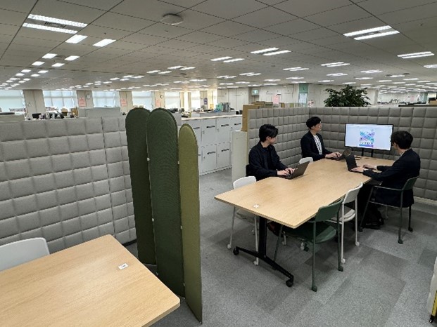 福祉局障害者施策推進部における、未来型オフィスの整備事例。長い机が手前と奥とでパーティションで区切って置かれており、奥の机には3人が座って打ち合わせをしている。