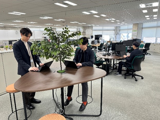 交通局建設工務部における、未来型オフィスの整備事例。手前にミーティング用のデスクが置かれており、2人が業務にあたっている。奥に個人執務用のデスクが置かれており、1人が座って業務にあたっている。